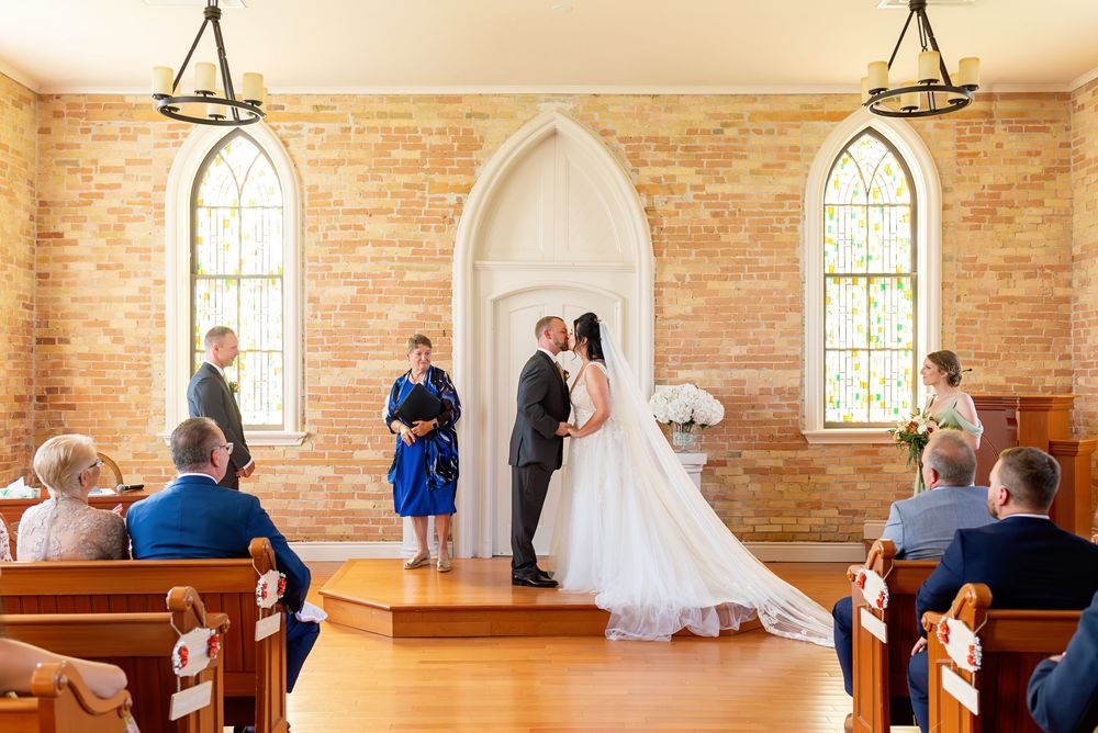 Bride and groom exchange vows at rustic brick chapel altar with arched windows and wedding guests seated in wooden pews.