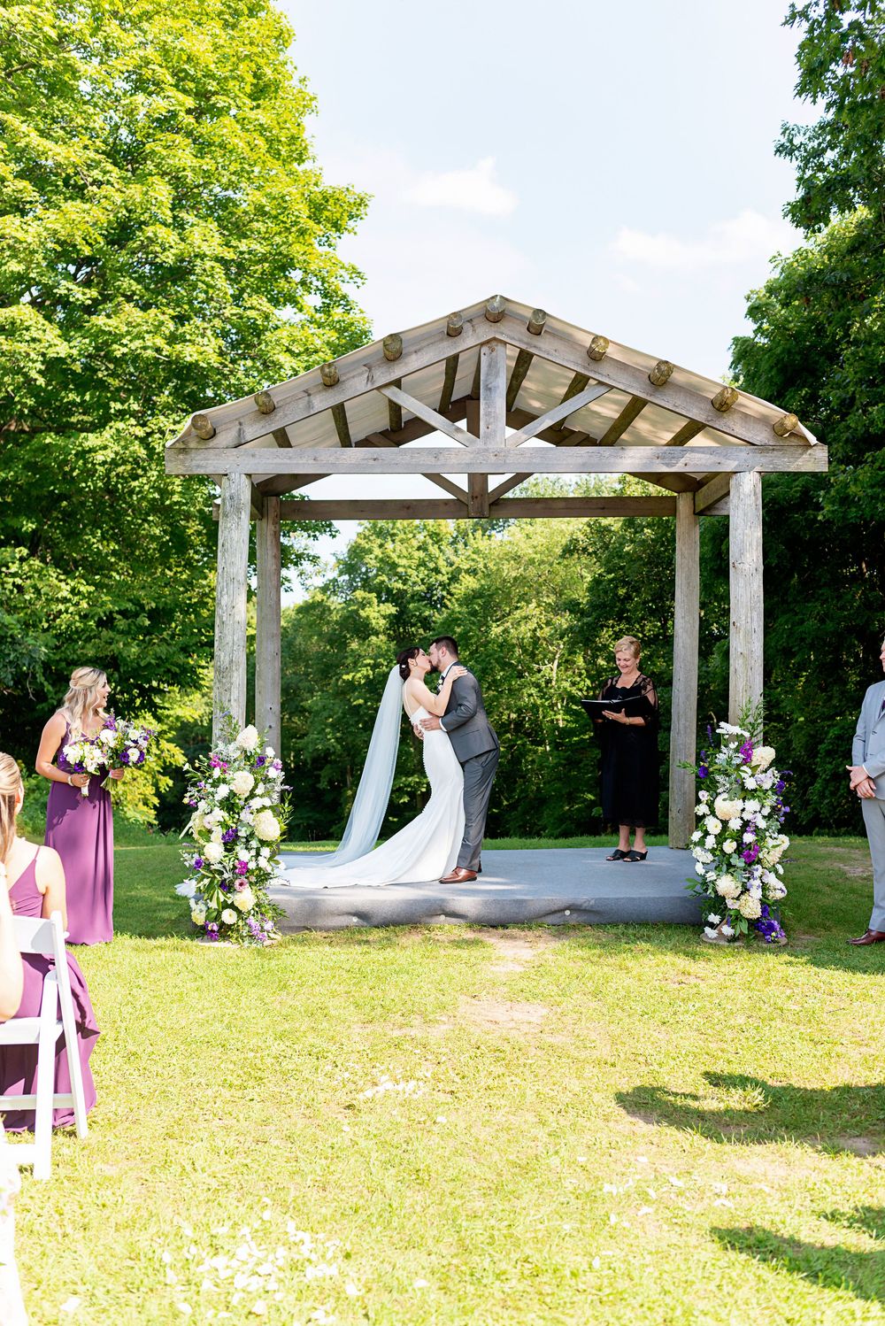 Couple shares first kiss under rustic wooden gazebo adorned with floral arrangements at outdoor wedding ceremony.