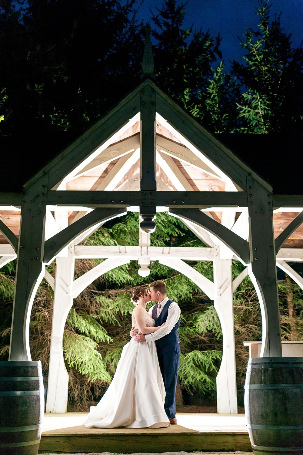 Bride and groom share a romantic kiss under an illuminated wooden arch gazebo at night, surrounded by lush evergreen trees.