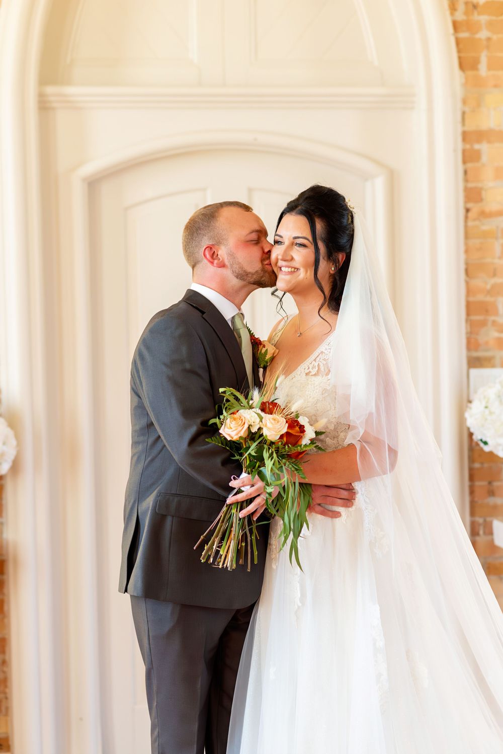Groom in gray suit kisses bride on cheek as she smiles holding a bouquet in a bright white wedding venue.