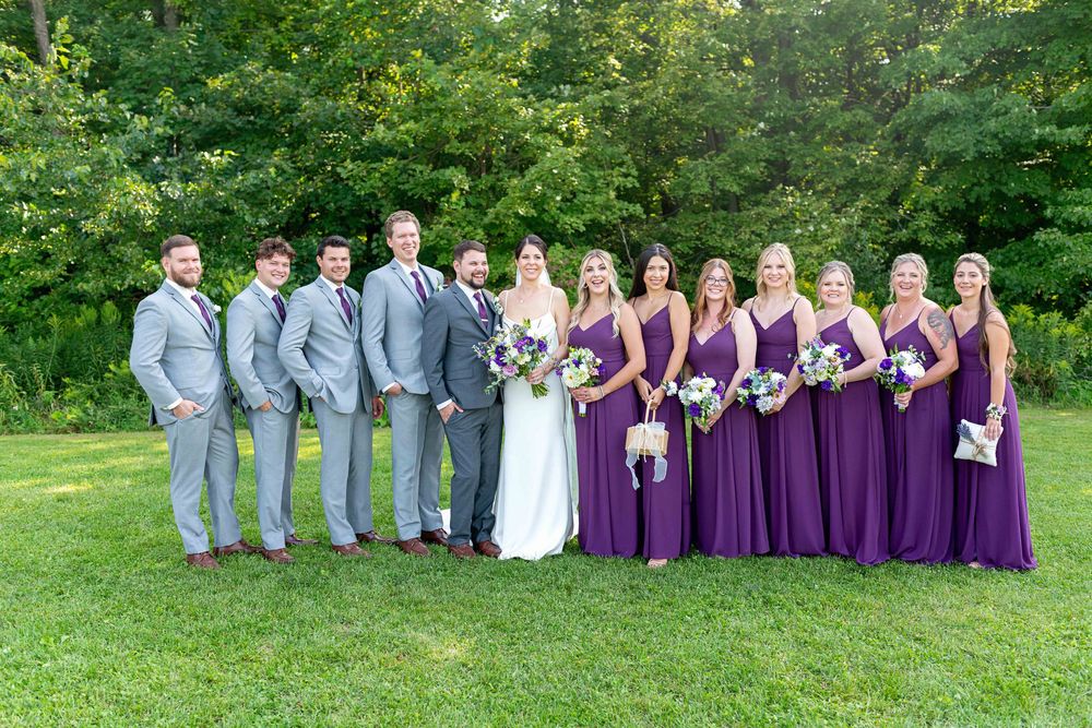 Wedding party posing outdoors, groomsmen in gray suits and bridesmaids in purple gowns flanking bride and groom on green lawn.