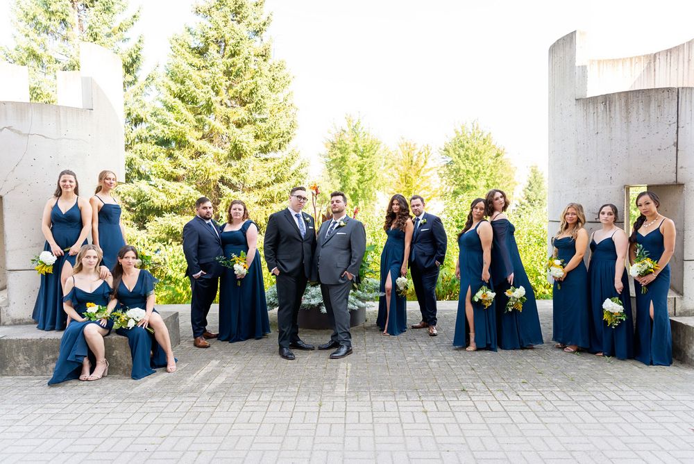 Wedding party in navy blue attire holding yellow bouquets posed outdoors at a modern venue with lush green trees.
