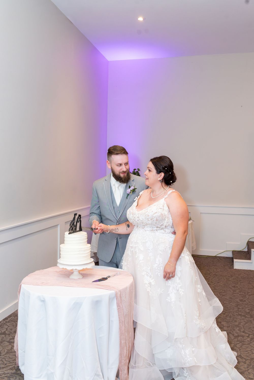 Bride and groom cutting their white wedding cake at reception, bride in lace gown, groom in gray suit, purple uplighting.