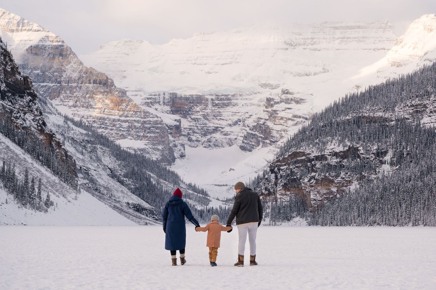 Family of three holding hands on frozen Lake Louise with snow-capped Rocky Mountains in the background.