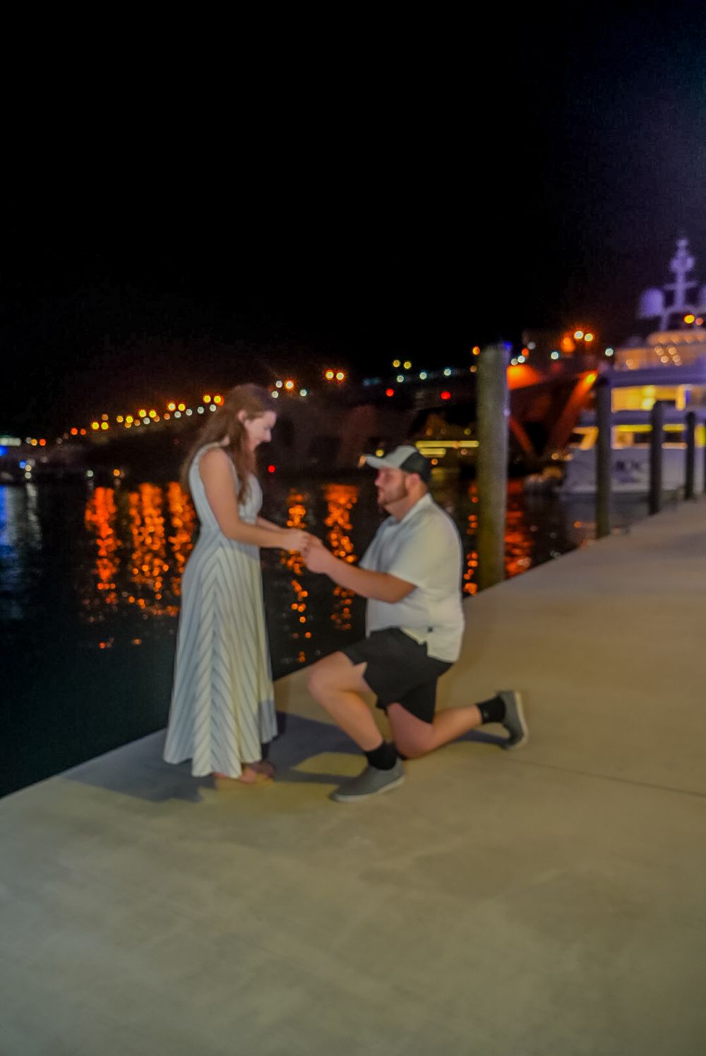 Man proposes to woman on one knee at a romantic waterfront dock at night with colorful lights reflecting on the water.