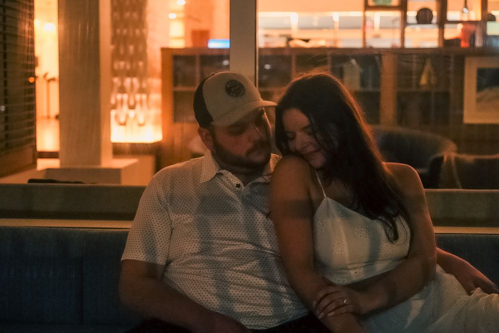Couple sitting close together on a couch in a warmly lit bar, woman resting her head on mans shoulder.