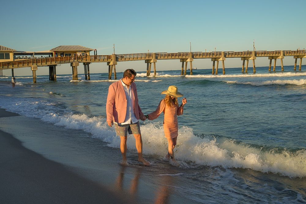 Couple holding hands walking along ocean shoreline near pier at golden hour.