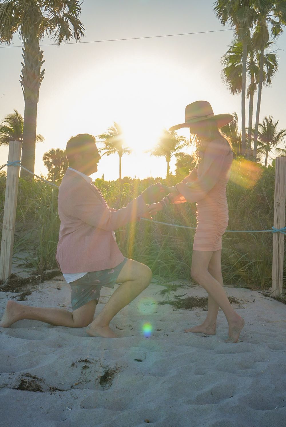Man proposing on one knee to woman on sandy beach at sunset with palm trees.