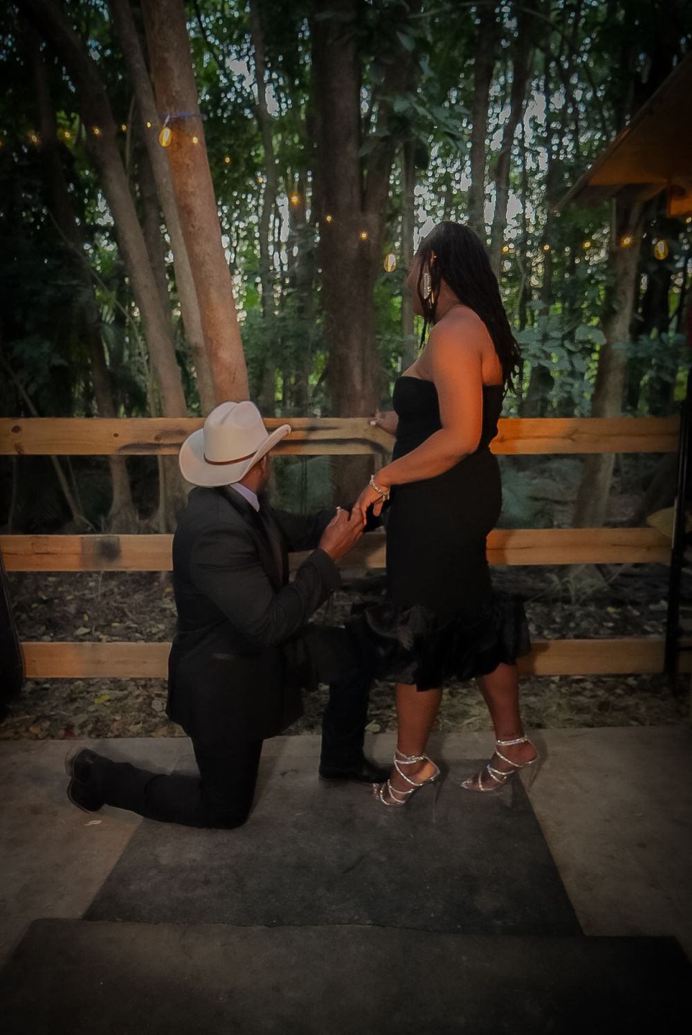 Man in cowboy hat kneeling on one knee proposing to woman in black dress outdoors at twilight.
