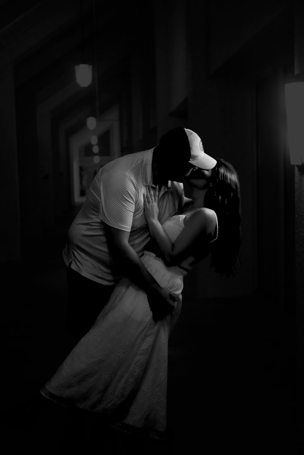 Black and white photo of a couple sharing a romantic kiss in a dimly lit hallway, man in cap dipping woman in white dress.