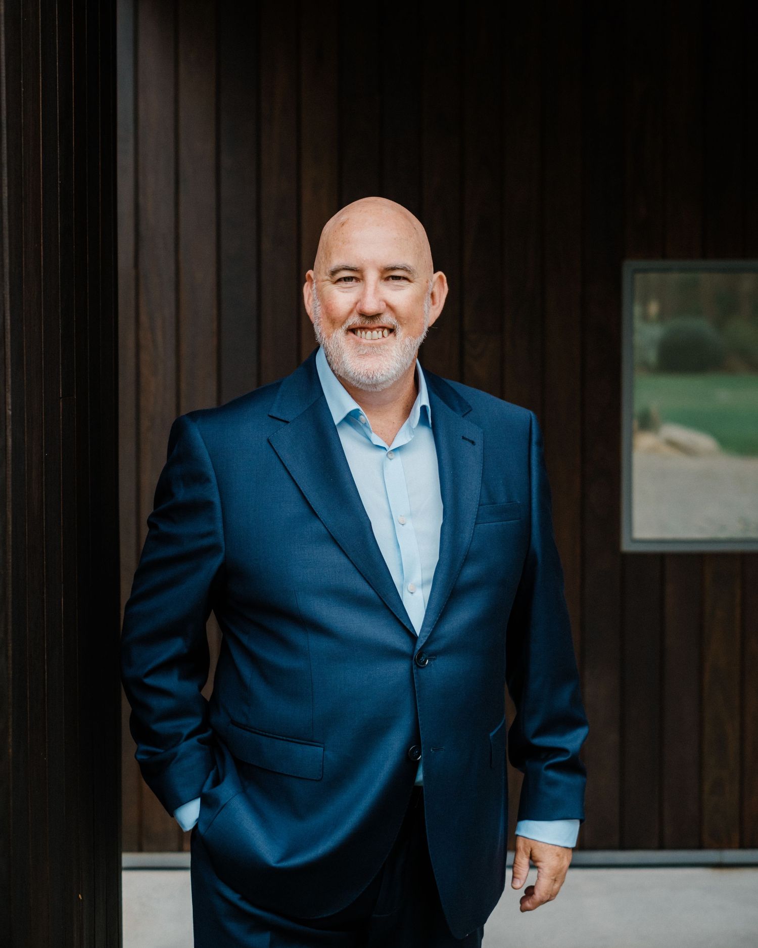 Smiling bald man in navy blue suit and light blue shirt standing confidently outdoors near modern wooden architecture.
