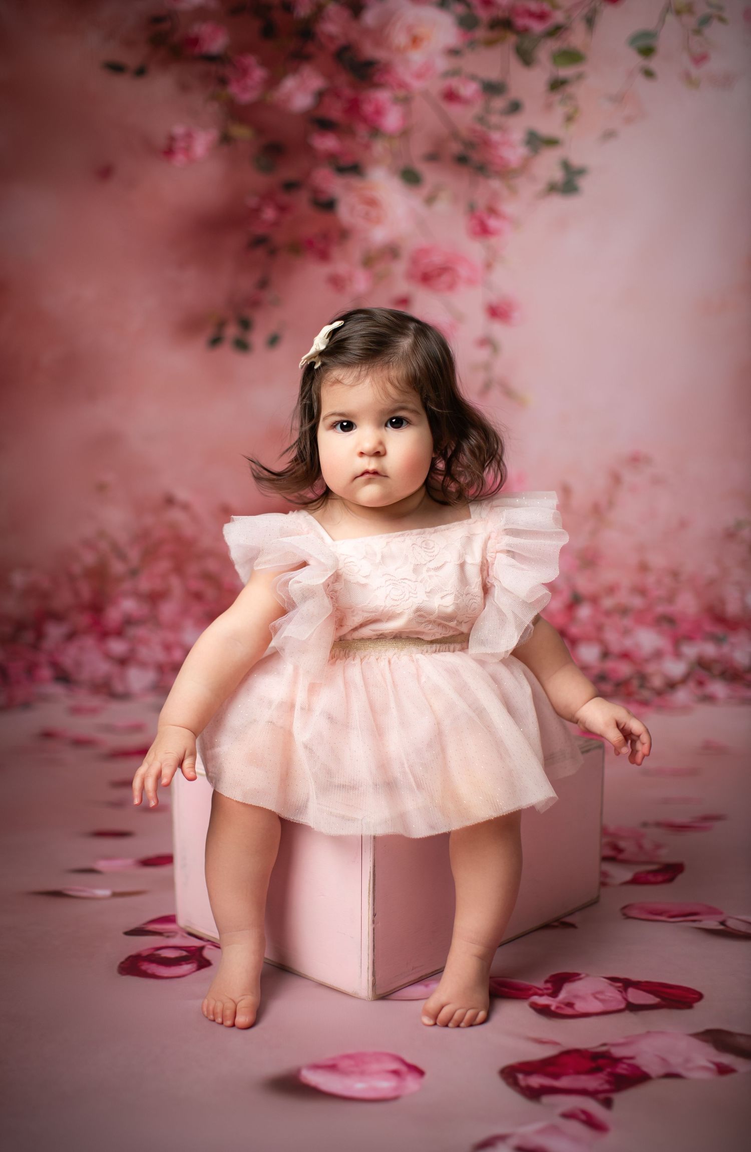 A baby girl in a purple floral tutu dress and headband sits on a lavender cube against a purple background.
