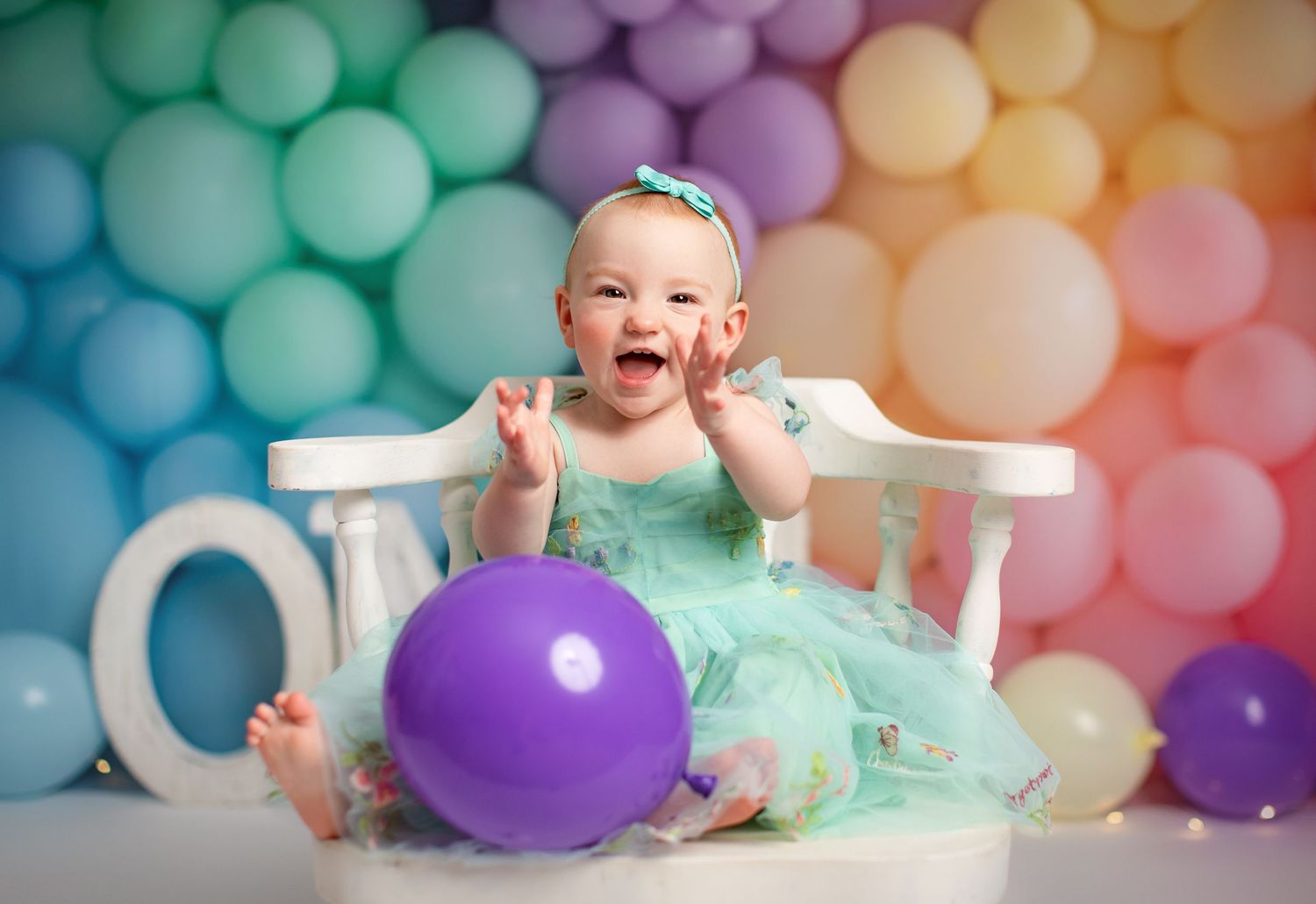 Baby girl in teal dress laughing in white chair surrounded by colorful rainbow balloons at first birthday party.