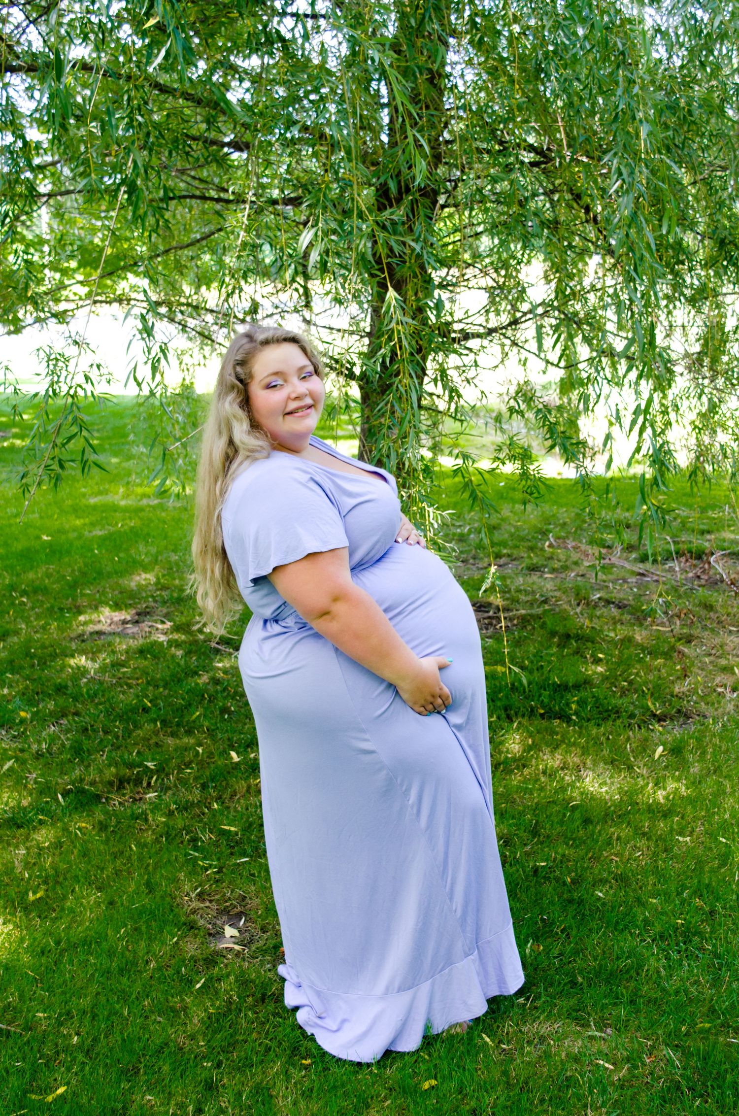 Pregnant woman in lavender maxi dress leaning on wooden bridge railing outdoors in lush green park setting.