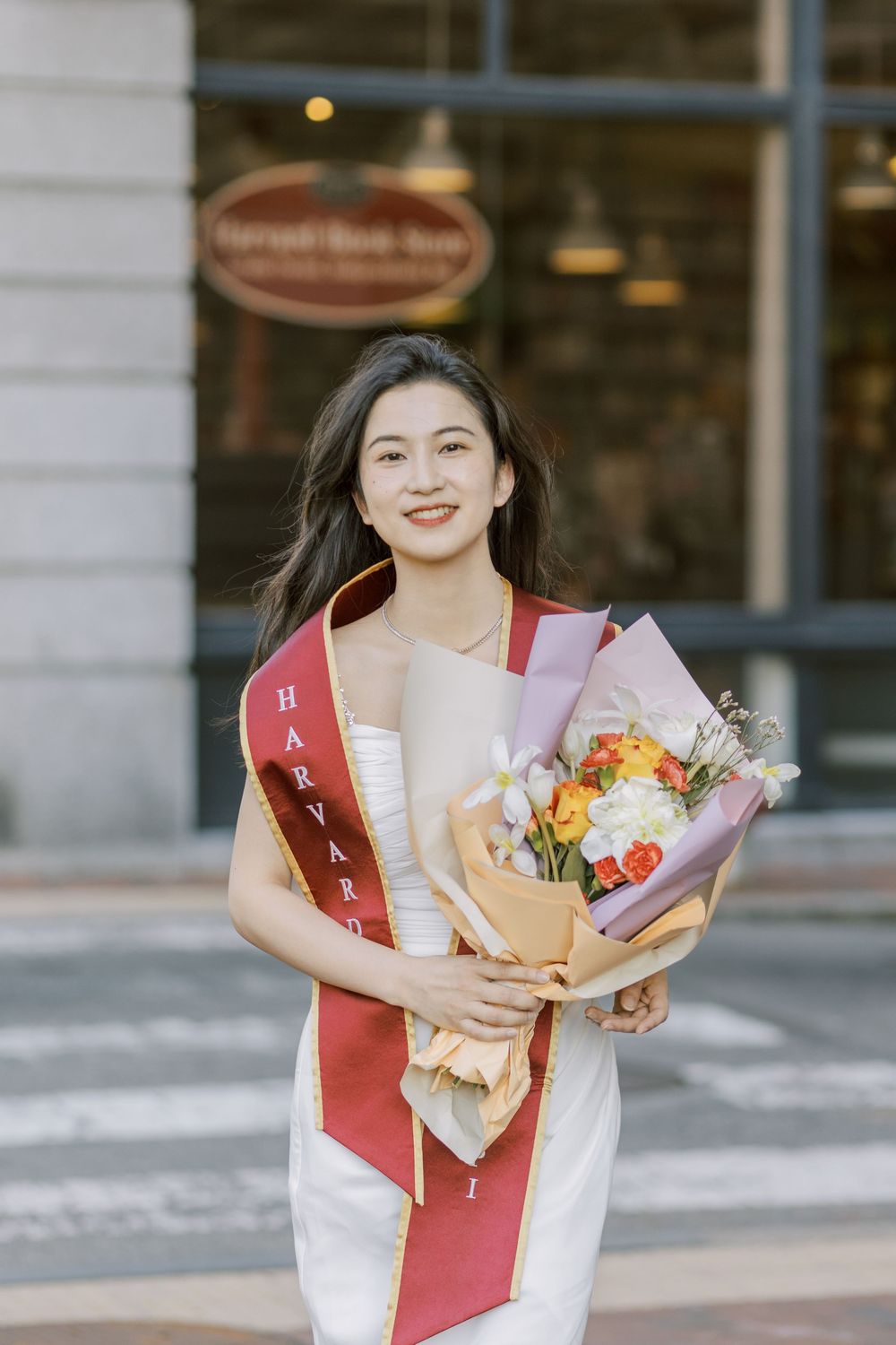 Young woman in white dress wearing Harvard sash holds colorful bouquet, smiling outdoors near building.