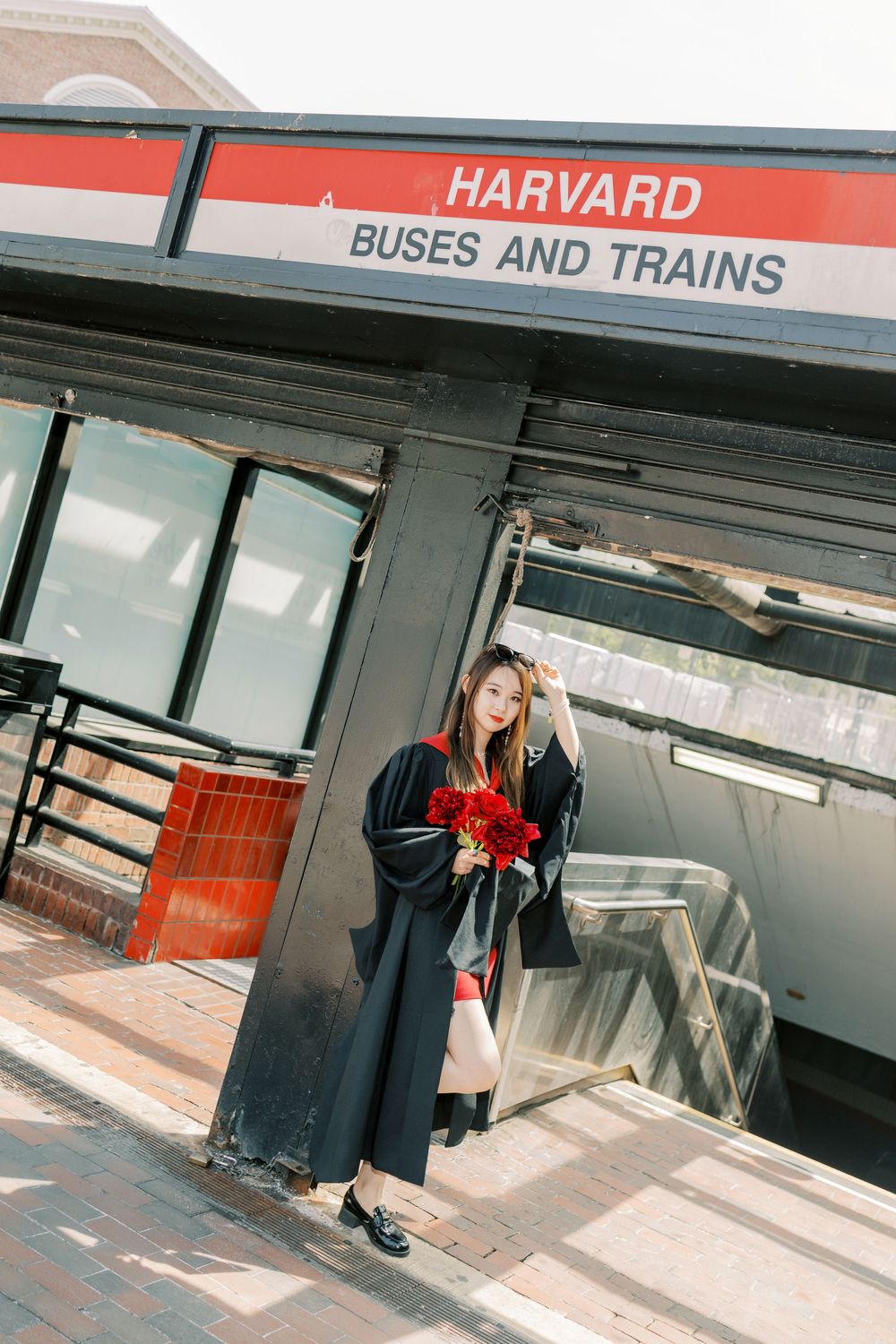 Harvard Graduate in cap and gown holds red roses at Harvard Buses and Trains station entrance.