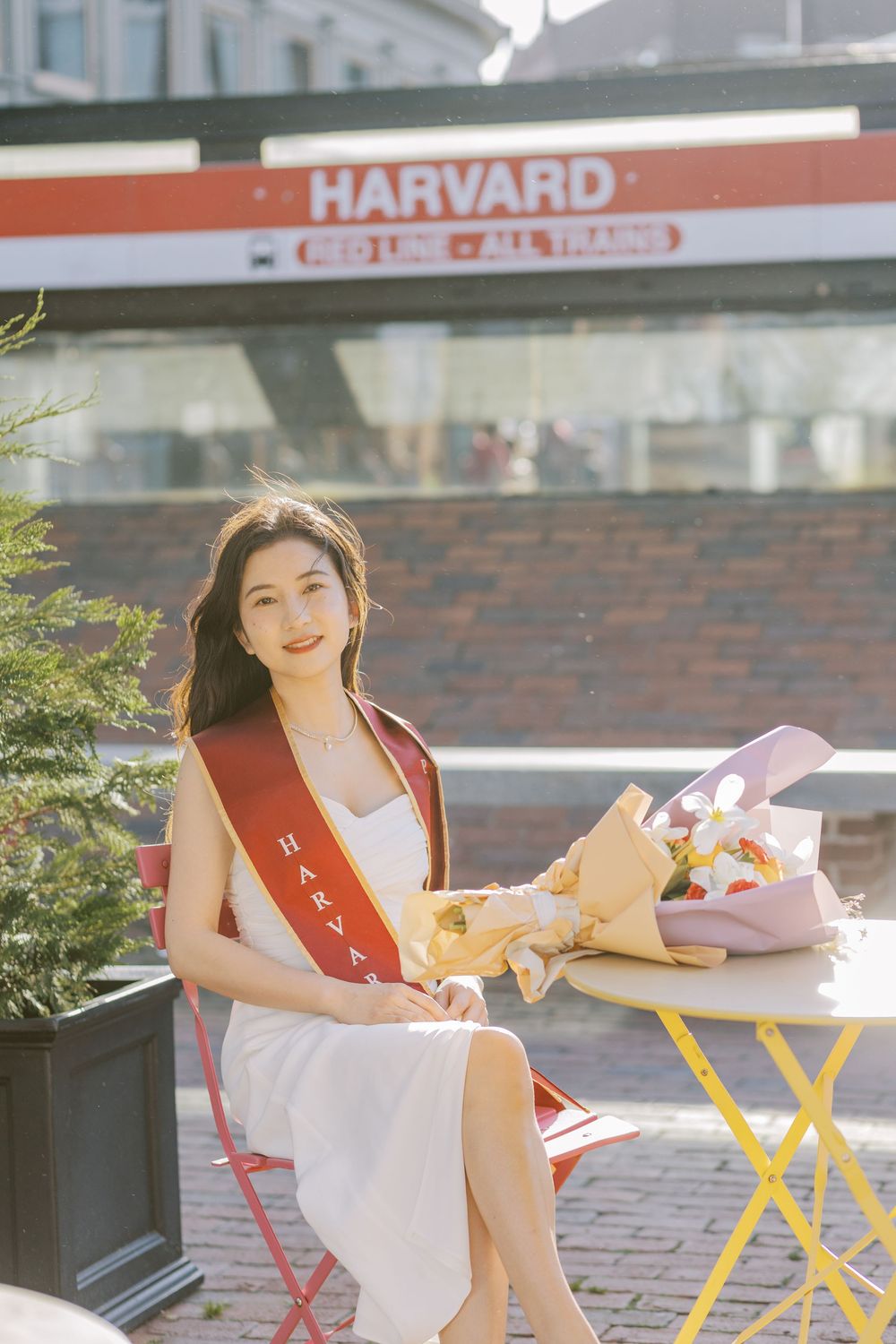 Young woman in white dress with Harvard graduation sash sits at outdoor table holding flowers near Harvard Square station.