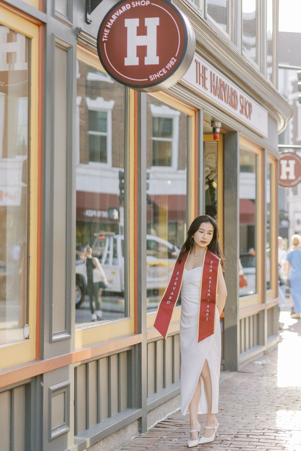 Harvard graduate in white dress and red sash poses proudly outside The Harvard Shop store on a sunny day.