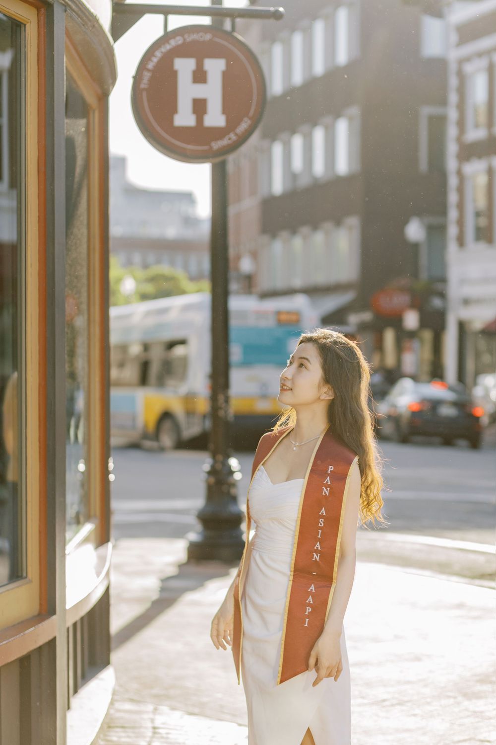 Young woman in white dress with red sash stands near Harvard sign on sunny urban street.