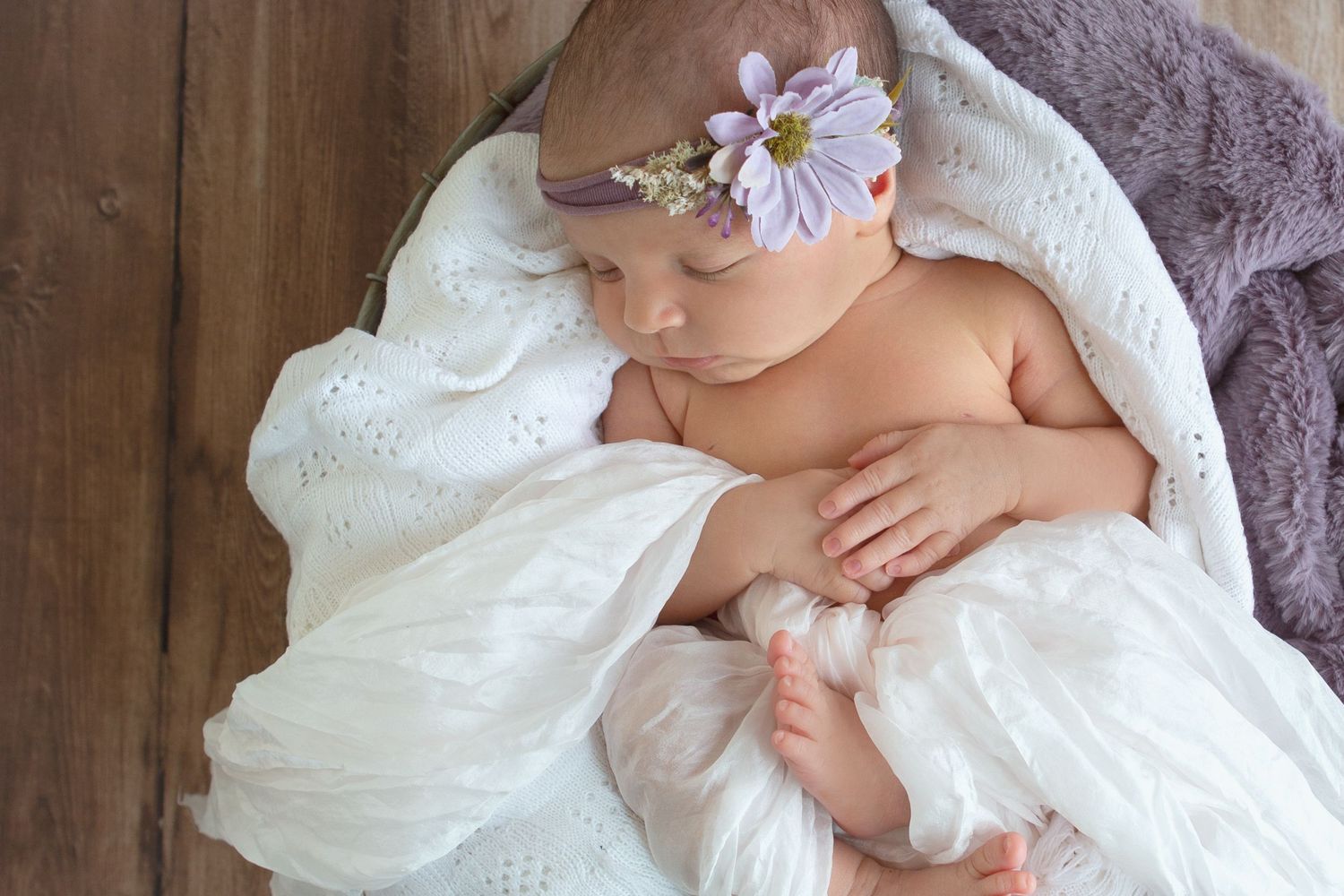 Newborn baby sleeping peacefully wrapped in white lace blanket wearing a purple floral headband on wooden background.