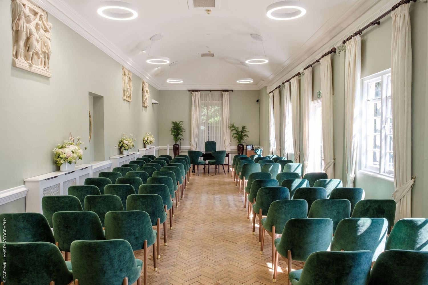 Elegant ceremony room with green velvet chairs arranged in rows, herringbone parquet flooring, and large windows with cream curtains.