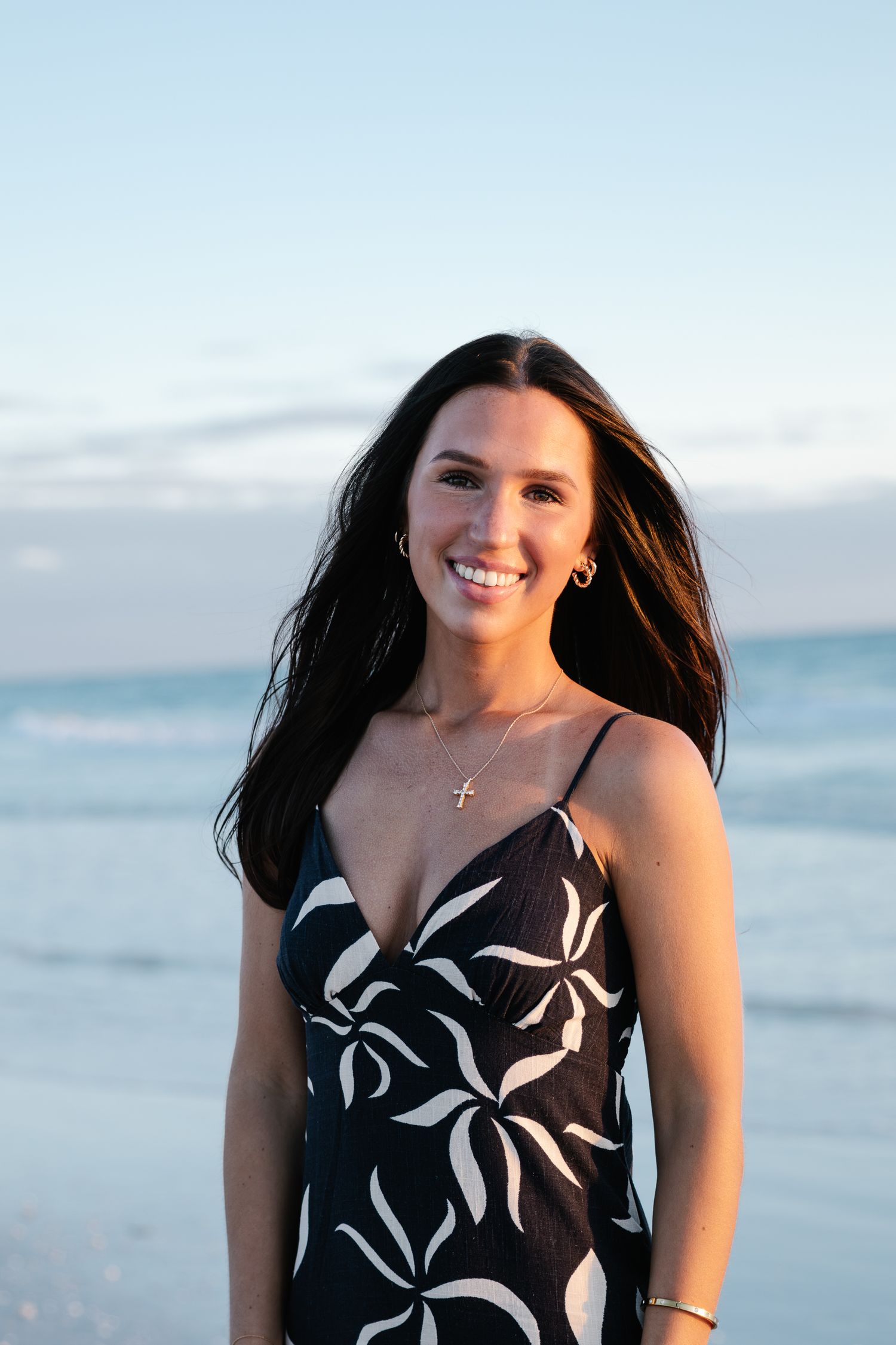 Young woman with long wavy hair poses at beach sunset in white top, hand in hair, ocean backdrop.