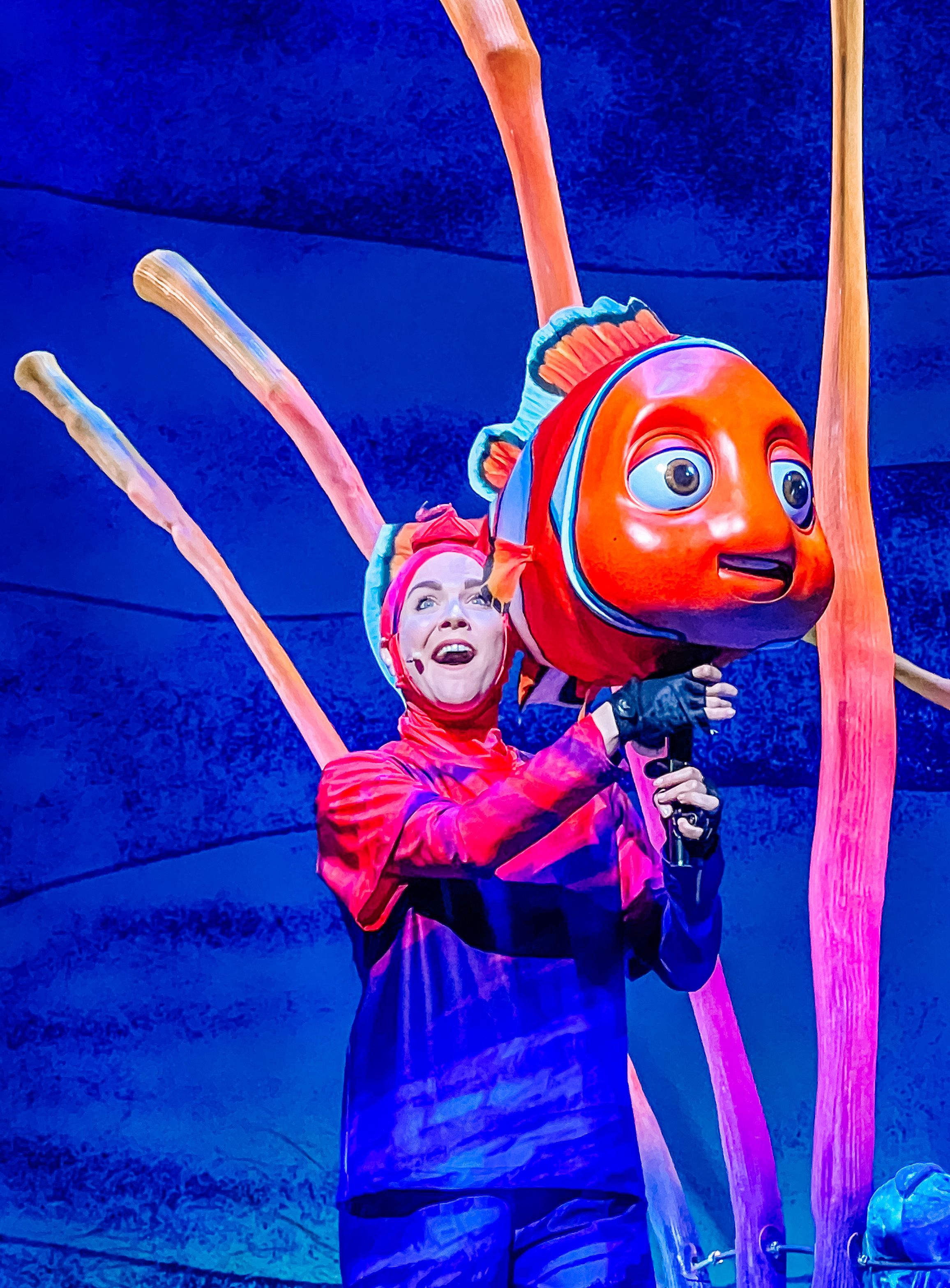 A puppet performer interacts with a bright orange Nemo-like fish character against a deep blue theatrical backdrop.