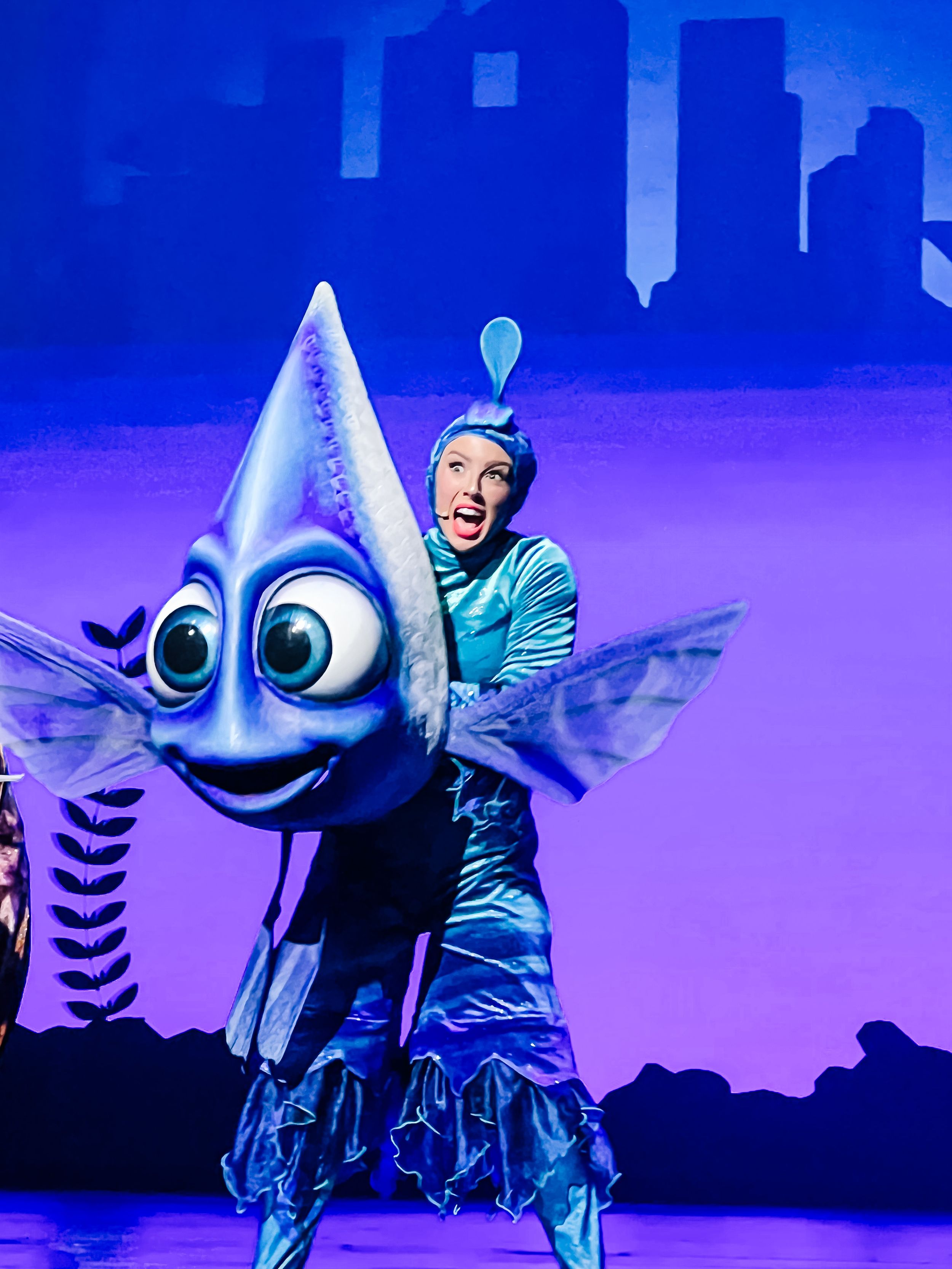 A performer in a blue costume stands next to a large smiling fish character puppet on a purple-lit stage set.