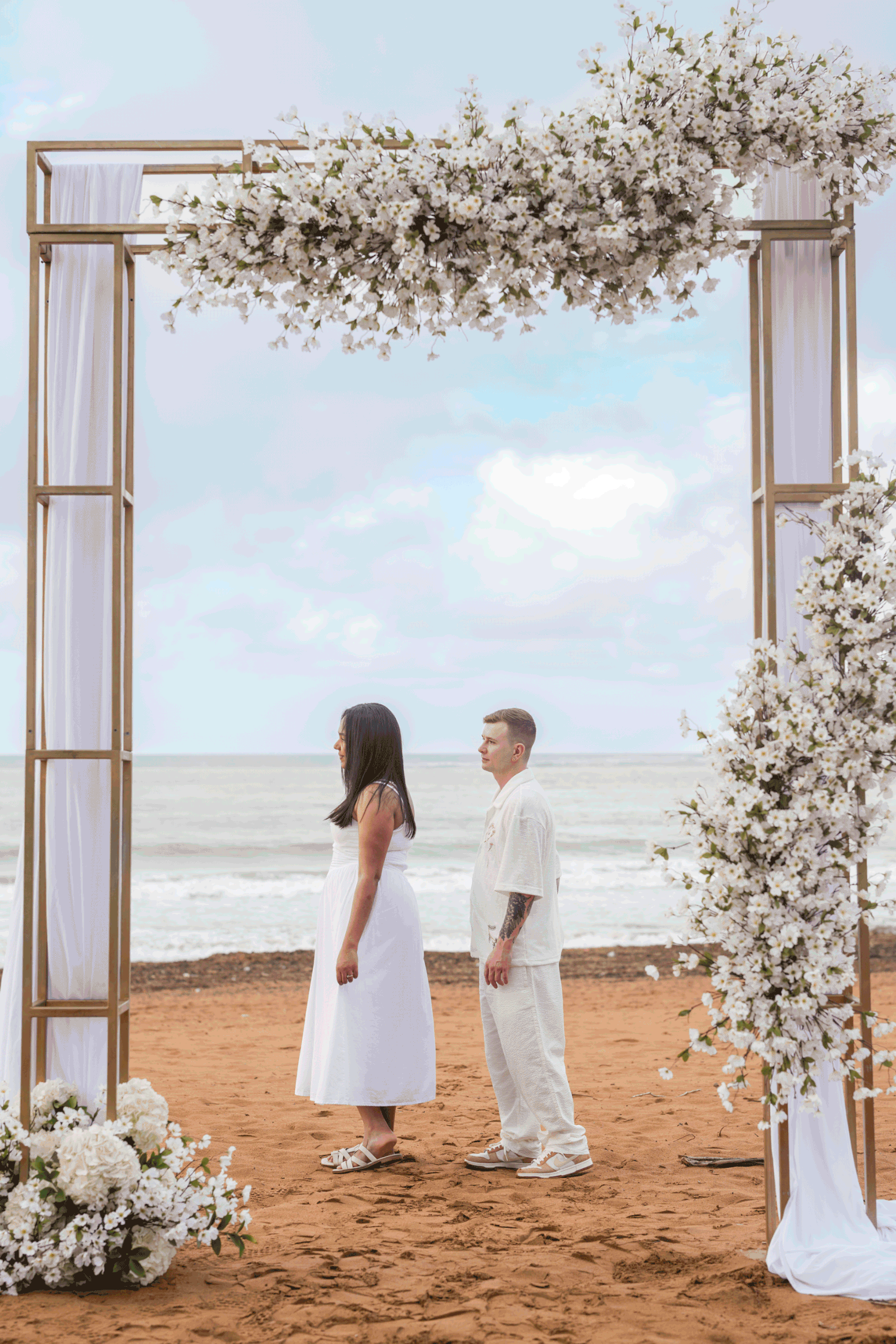 •	Bride sitting under a floral arch by the ocean during wedding ceremony
