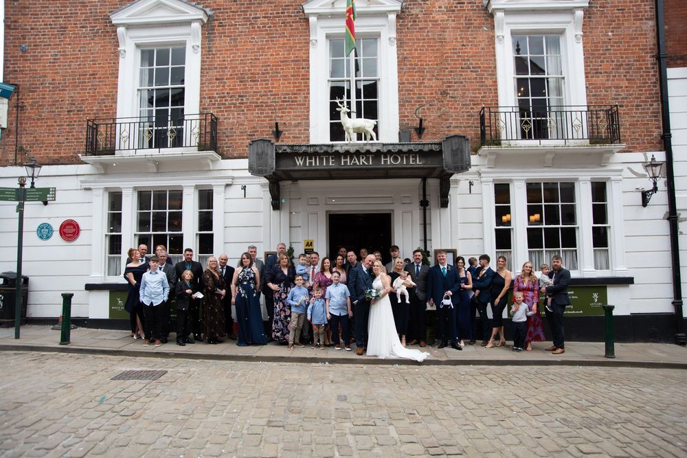 Wedding party posing outside the White Hart Hotel, a historic brick building, with bride and groom at center.