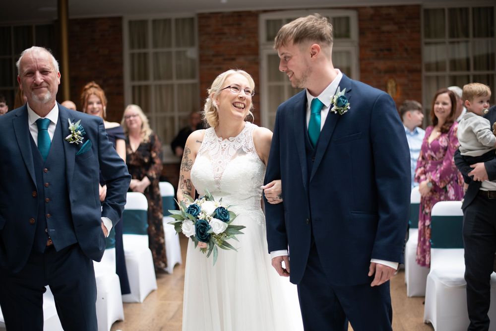 Bride and groom walking down the aisle smiling, bride holding teal bouquet, guests seated in decorated wedding venue.