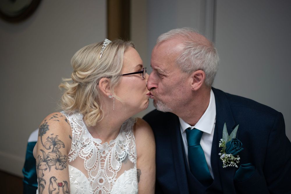 Bride and groom sharing a kiss at their wedding ceremony, bride wearing lace dress with tattoos visible.