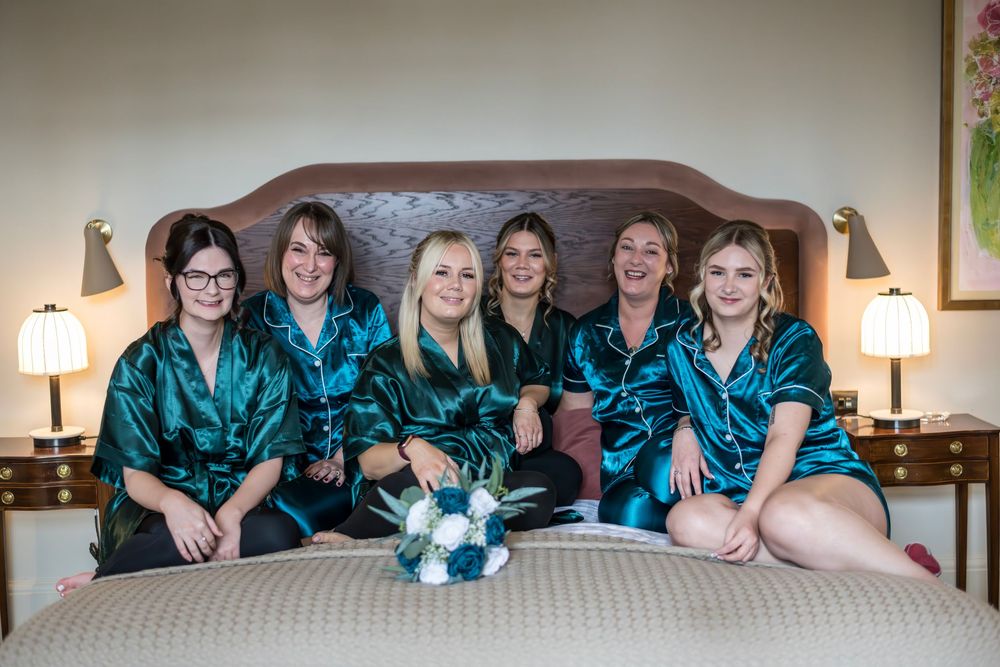 Six bridesmaids in matching teal satin robes sitting on a hotel bed with a bridal bouquet during wedding preparations.