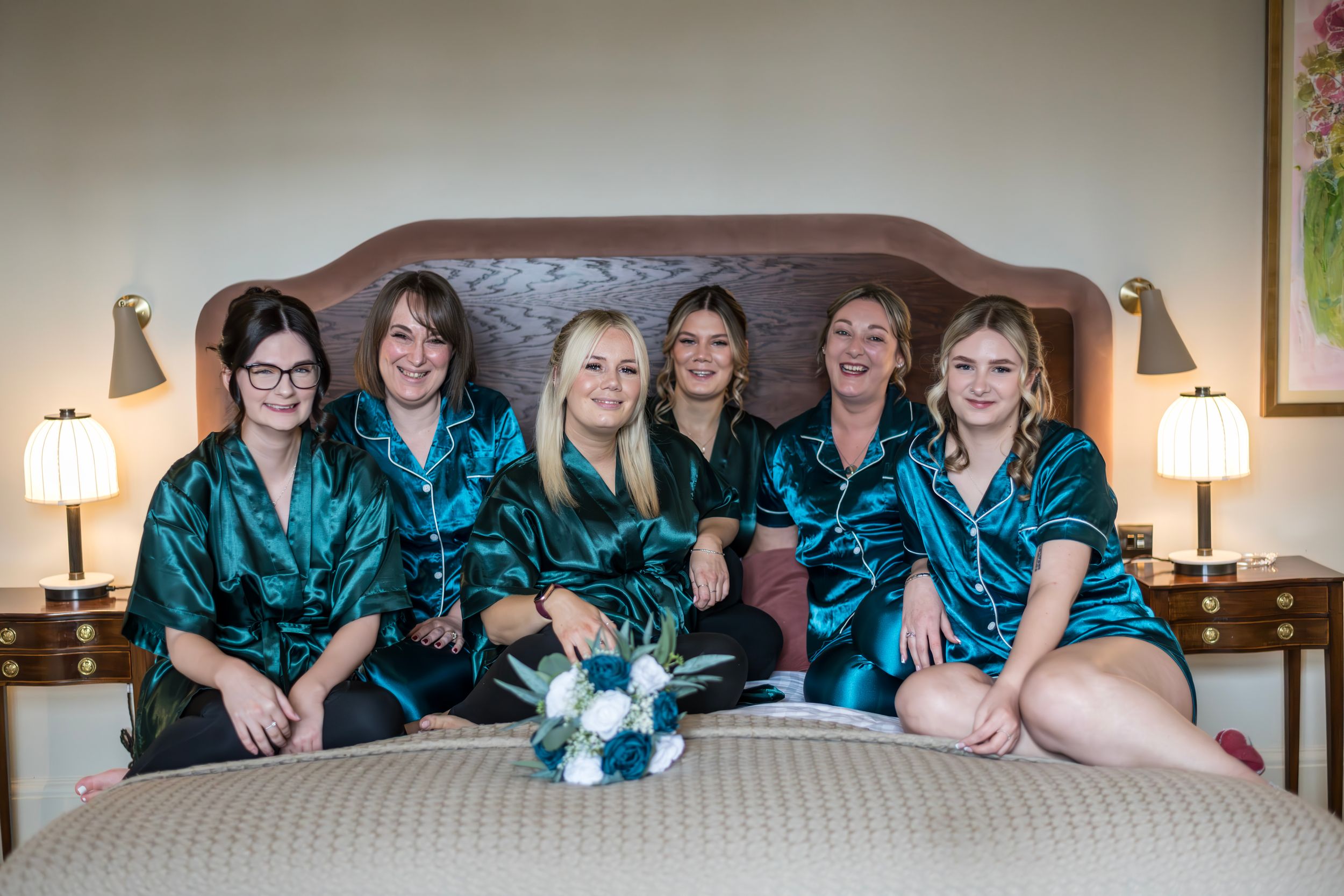Six bridesmaids in matching teal satin robes sitting on a hotel bed with a bridal bouquet during wedding preparations.
