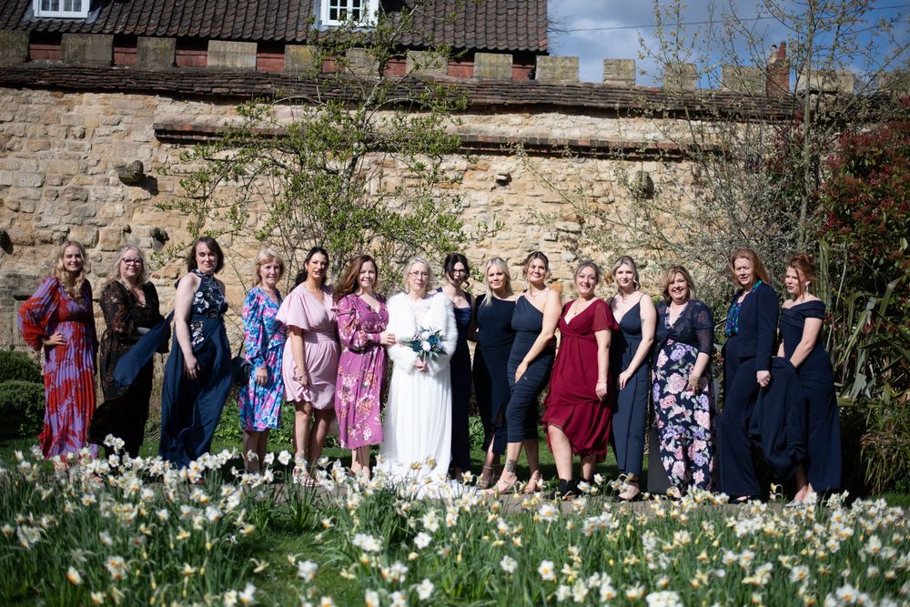 Bride and bridesmaids posing together in a garden with blooming daffodils in front of a stone wall.