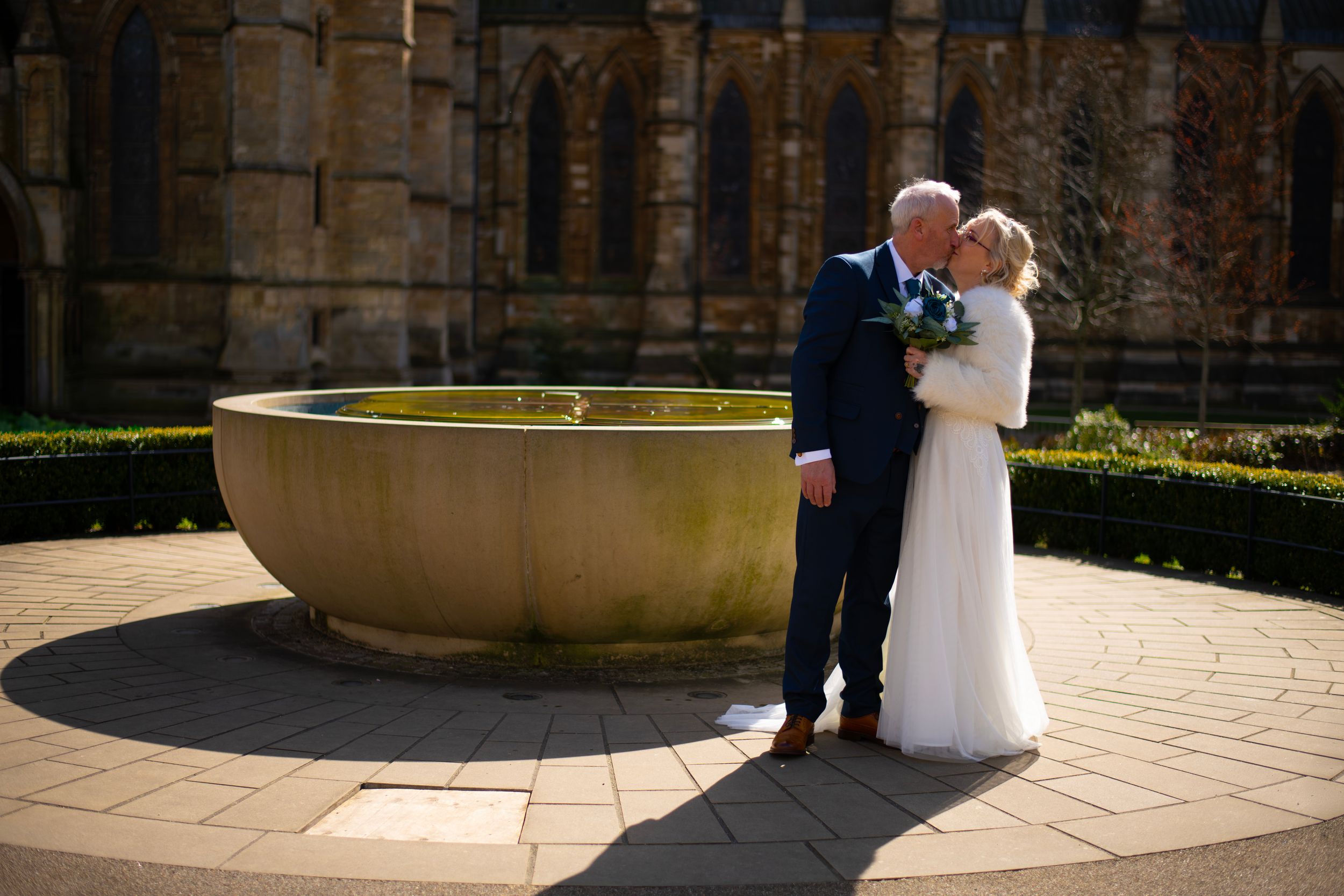 Bride and groom share a romantic kiss beside a stone fountain outside a historic church on their wedding day.