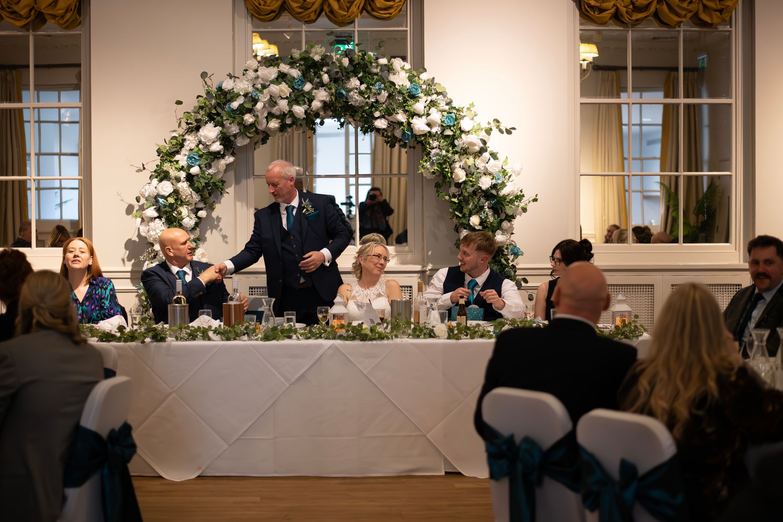 Elegant wedding reception with floral arch, head table, and guests listening to a speech in a grand ballroom.