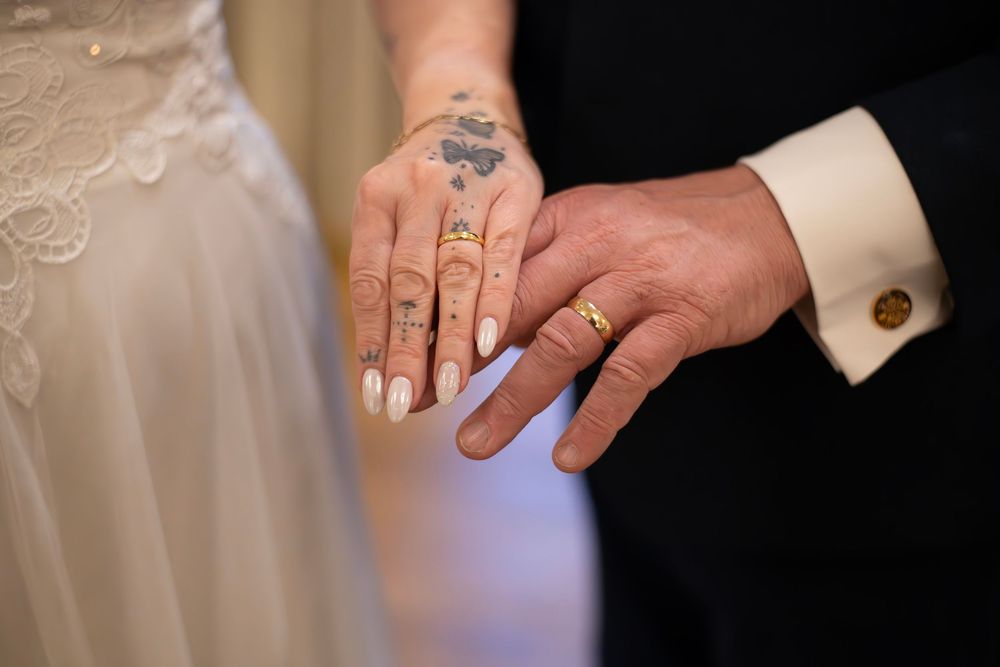 Close-up of wedding couple exchanging rings, bride with tattooed hand and white nails, groom wearing gold band.