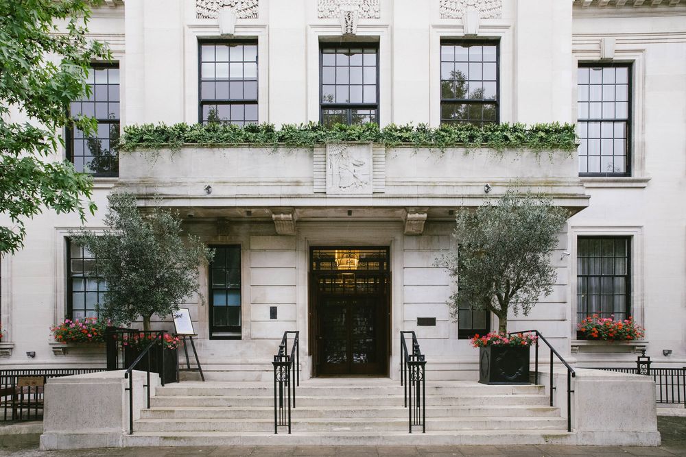 Elegant white neoclassical building entrance with stone steps, potted olive trees, and lush rooftop greenery in London.