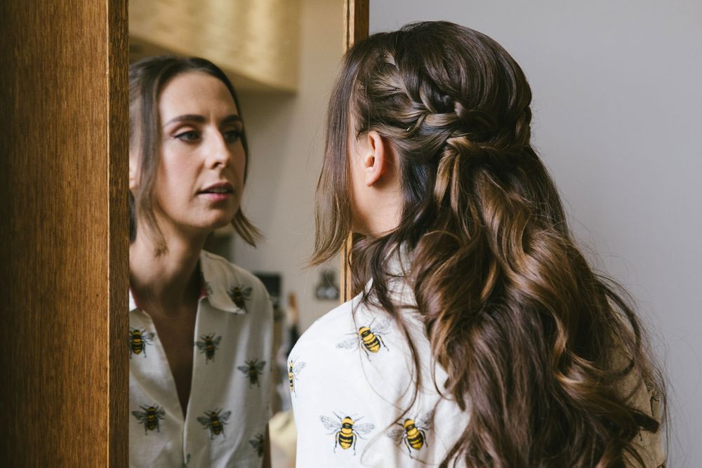 Woman with braided half-up hairstyle wearing bee-print blouse gazes at her reflection in a wooden-framed mirror.
