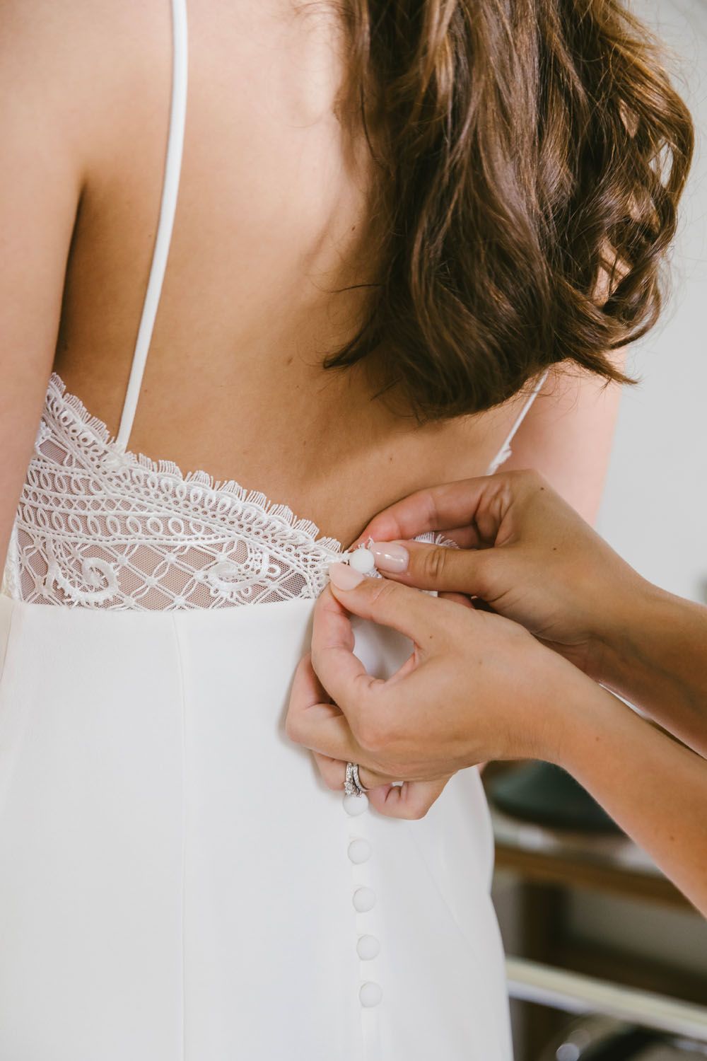 Helper fastening buttons on back of white lace wedding dress for bride getting ready.
