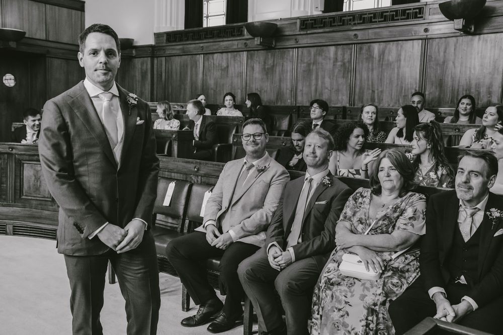 Groom stands at altar in suit while wedding guests smile in church pews during ceremony.