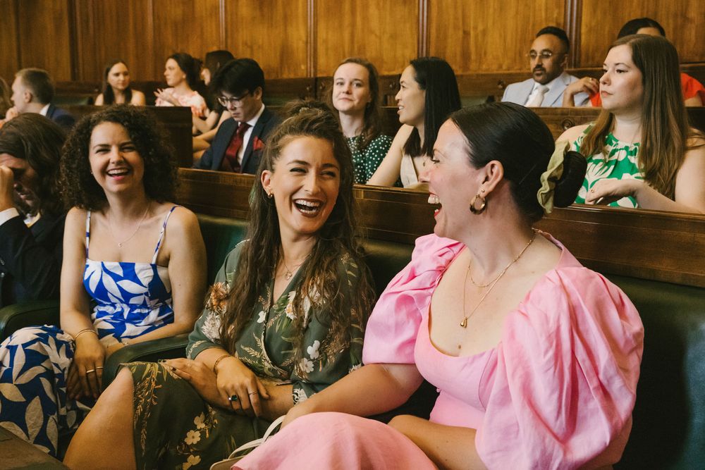 Three women laughing joyfully while seated in wooden pews at a formal event or ceremony.