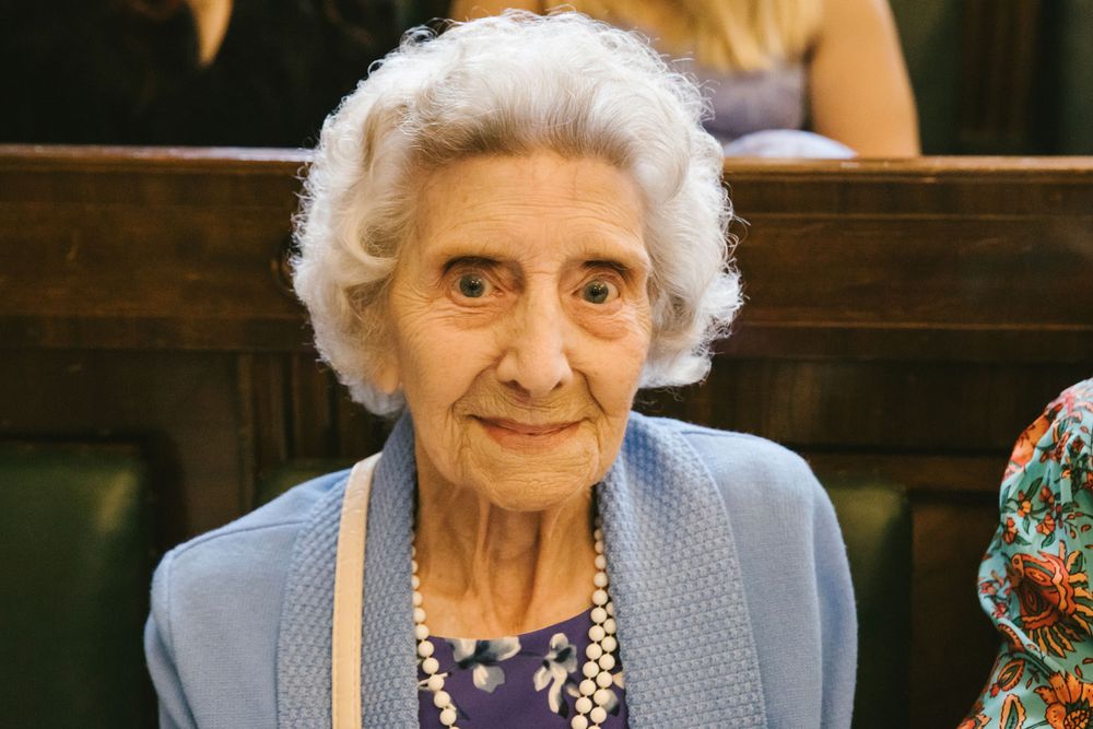 Elderly woman with white hair and pearl necklace smiling in church pew at wedding ceremony.