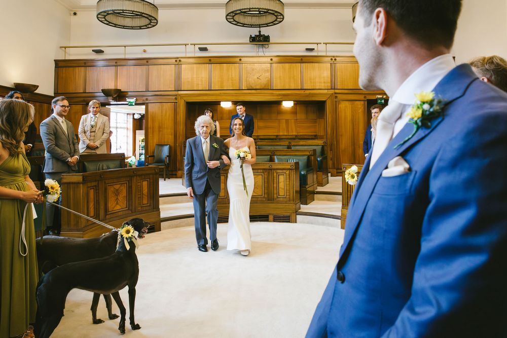 Bride walks down the aisle with her father at a registry office wedding ceremony, with a black greyhound dog in attendance.