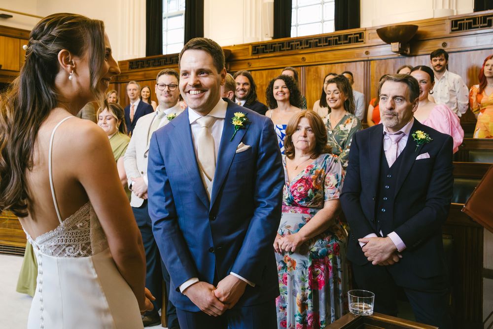Groom in blue suit smiling at bride during wedding ceremony with guests watching in elegant wood-paneled venue.