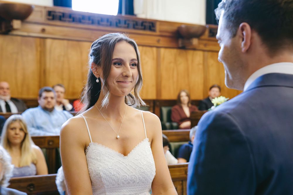 Bride in white dress smiling at groom during indoor wedding ceremony with guests seated in wooden pews.