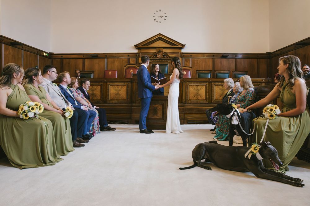 Elegant wedding ceremony in a wood-paneled hall with bridesmaids in olive green dresses and a dog sitting beside seated guests.