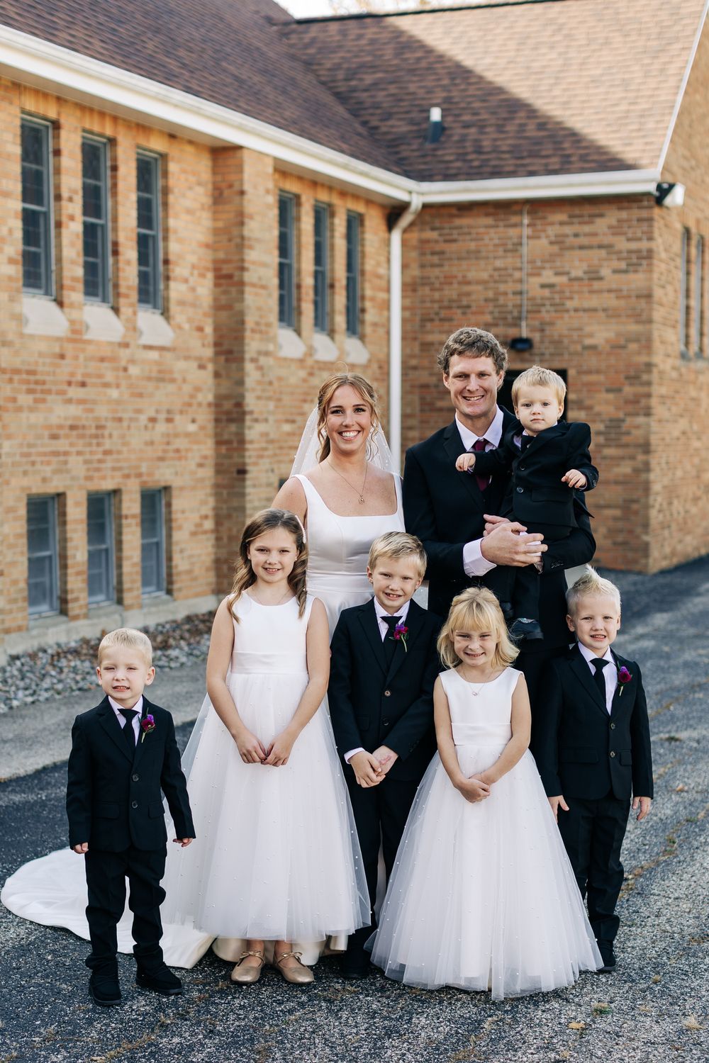 Bride and groom pose with flower girls and ring bearers in formal attire outside a brick church.