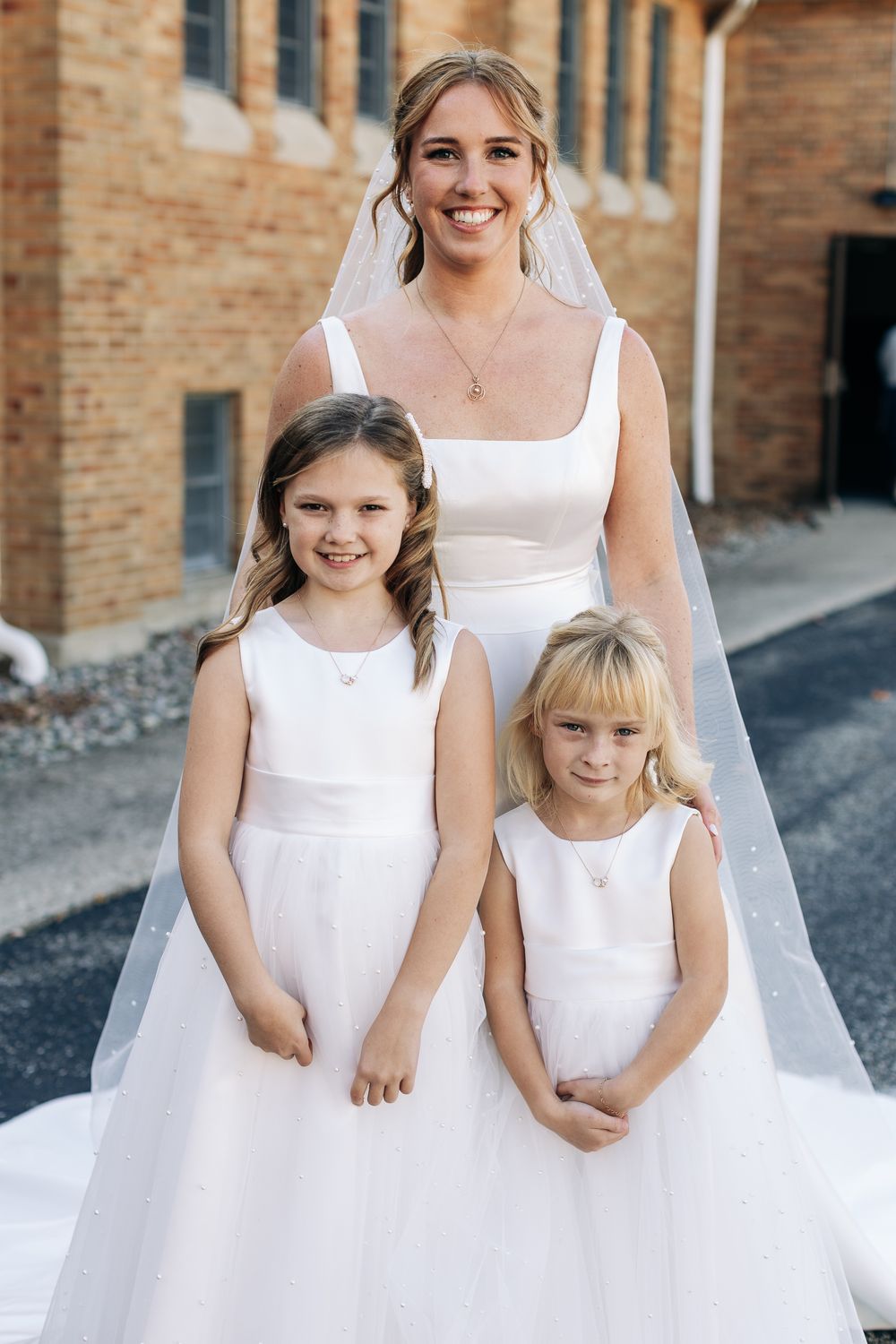 Smiling bride in white gown and veil poses with two flower girls in white dresses outside a brick building.